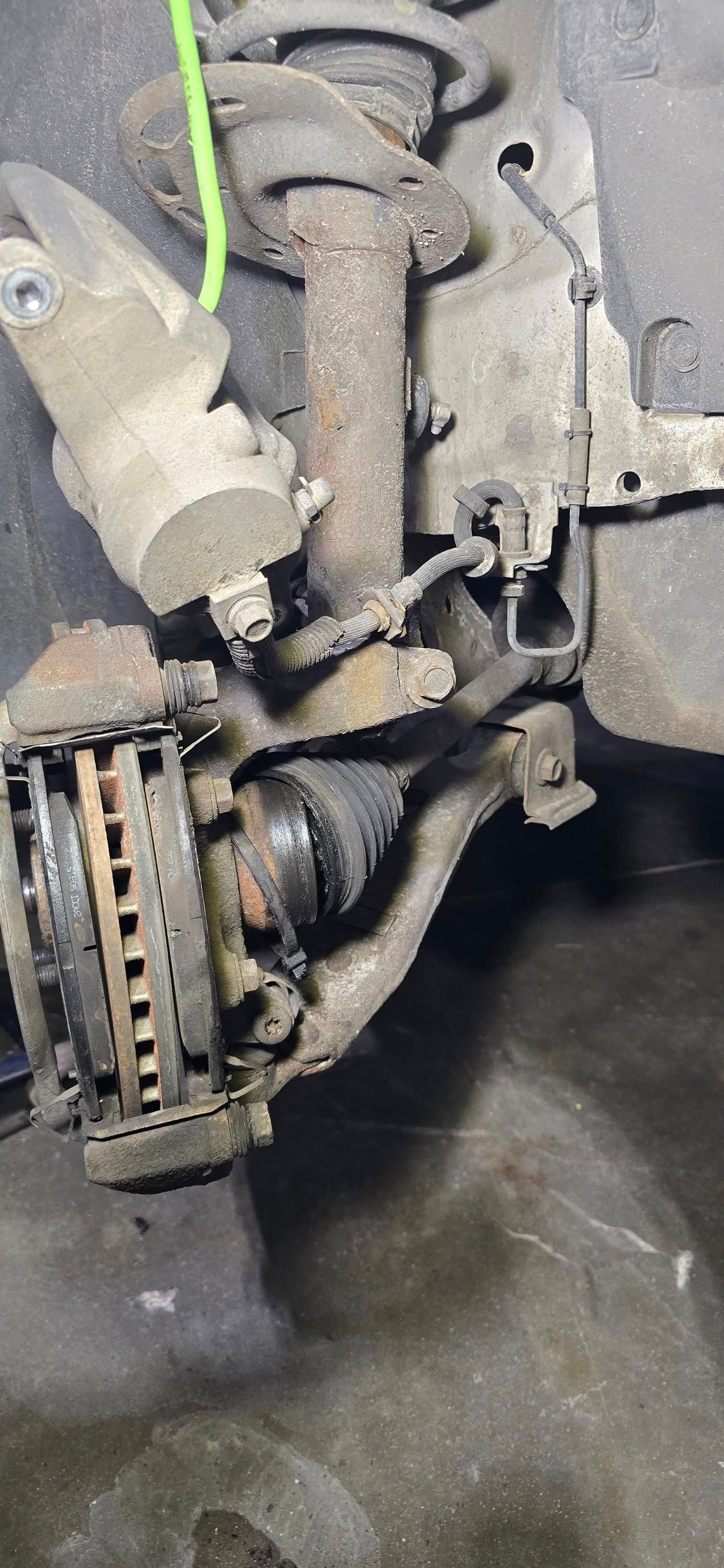 Close-up of a car brake rotor, caliper, and suspension components with a green hose attached, showing heavy wear.