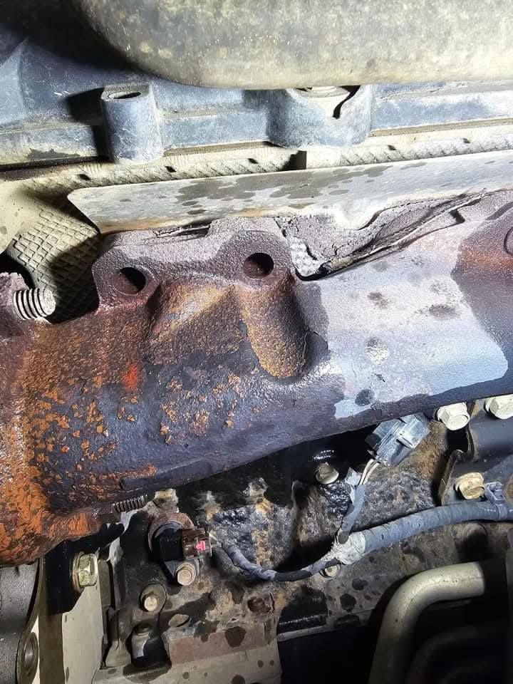Rusty exhaust manifold and engine components under a car hood, with blackened heat damage.
