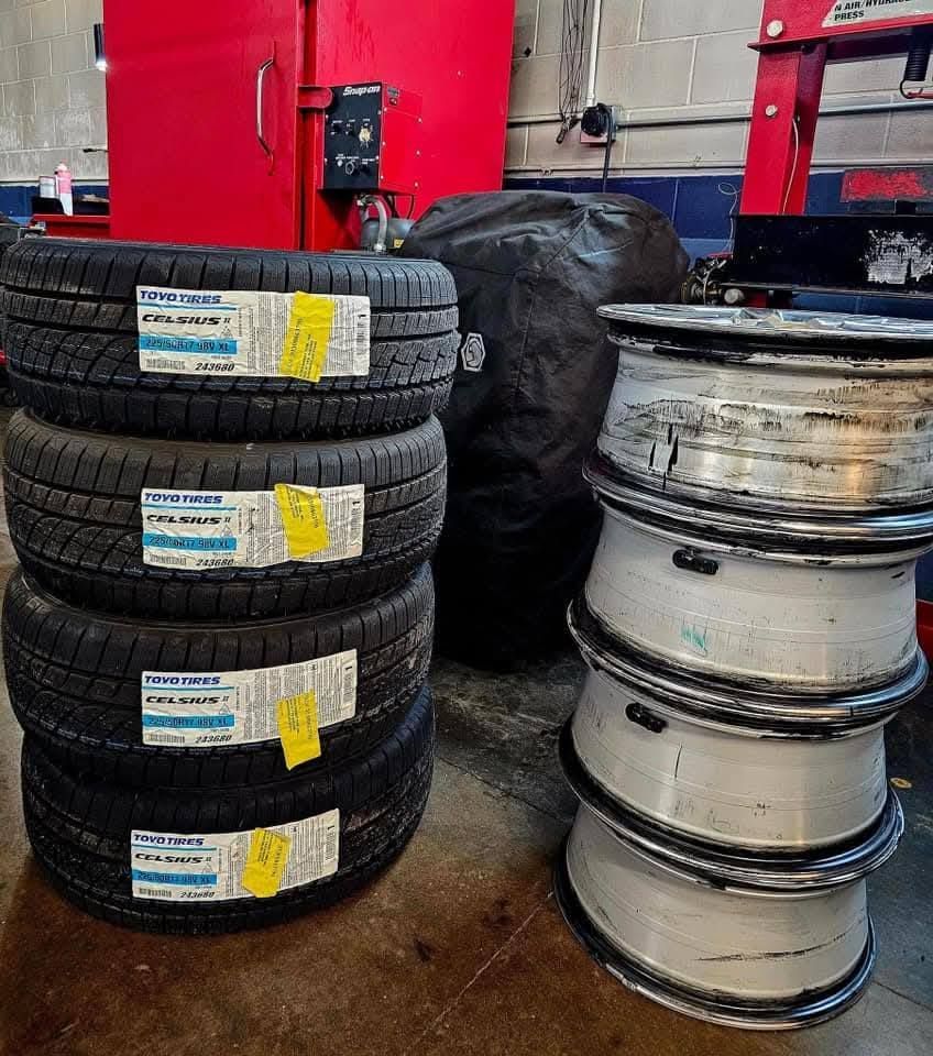 Stacks of truck tires and silver rims in a garage workshop, with red equipment in the background