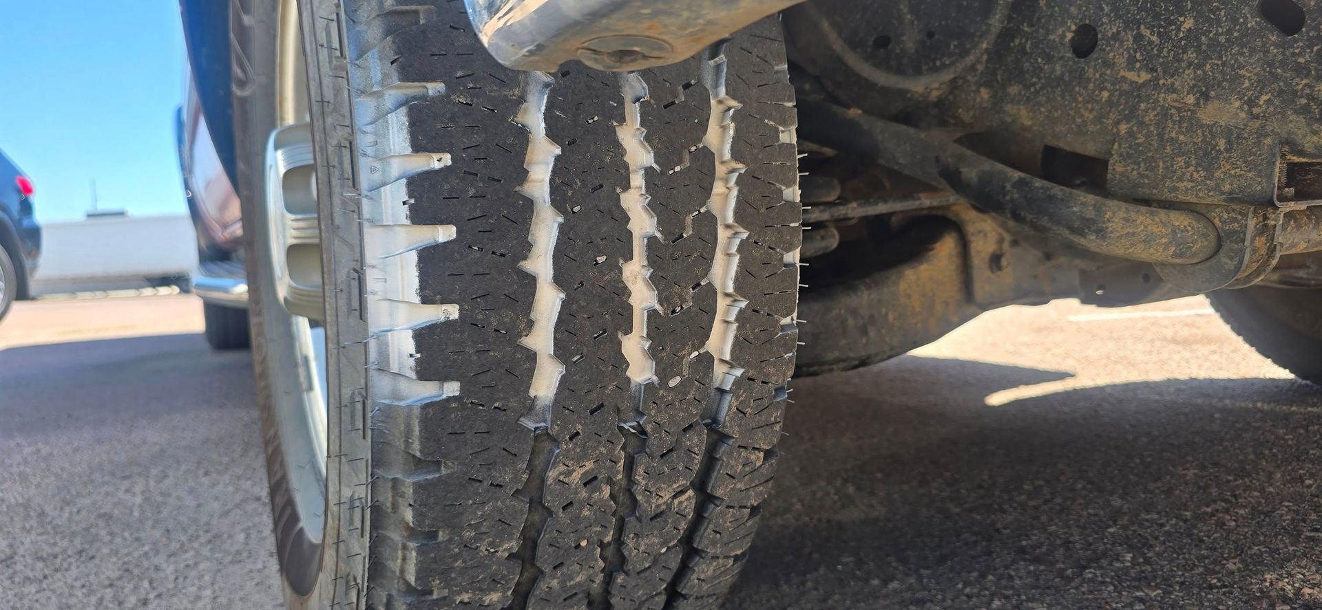 Close-up of a car tire with worn tread on a sunny road, beside another vehicle in the background