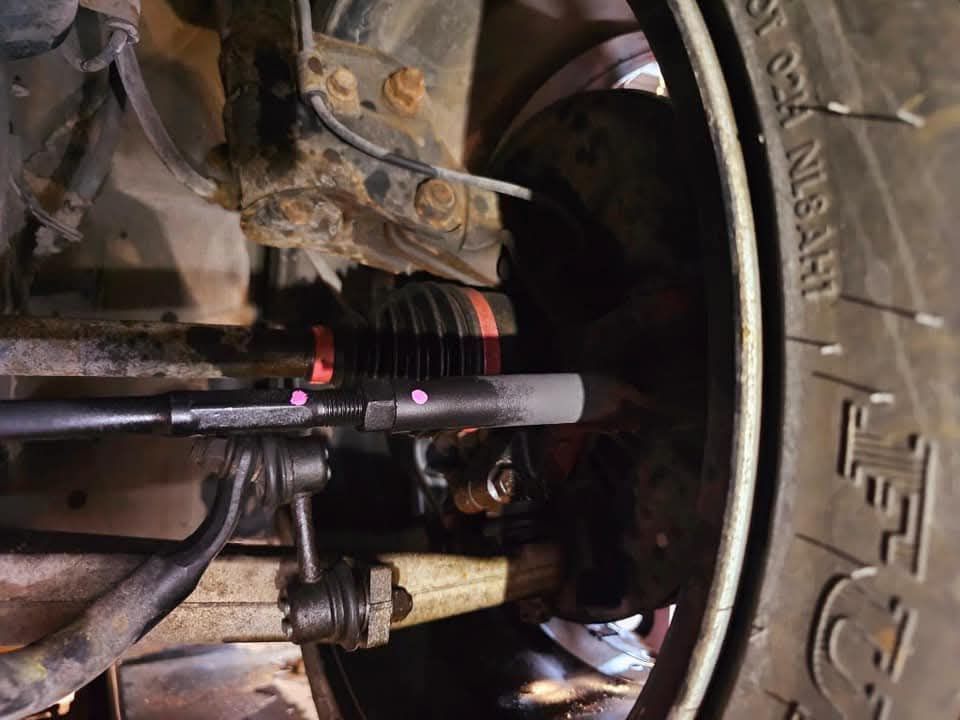 Close-up of a vehicle undercarriage with suspension parts and a mounted black cylindrical rod with pink tape markers.