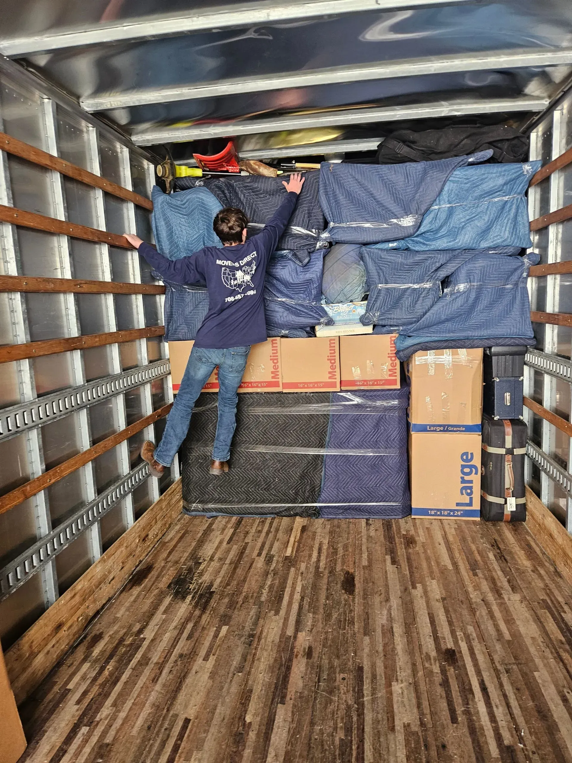 Person in a moving truck, arms outstretched, standing on stacked furniture and boxes. Truck interior is visible.