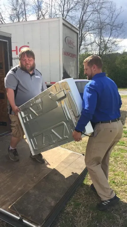 Two men carrying a large, white appliance, likely a refrigerator, onto a moving truck. Outdoors, sunny.