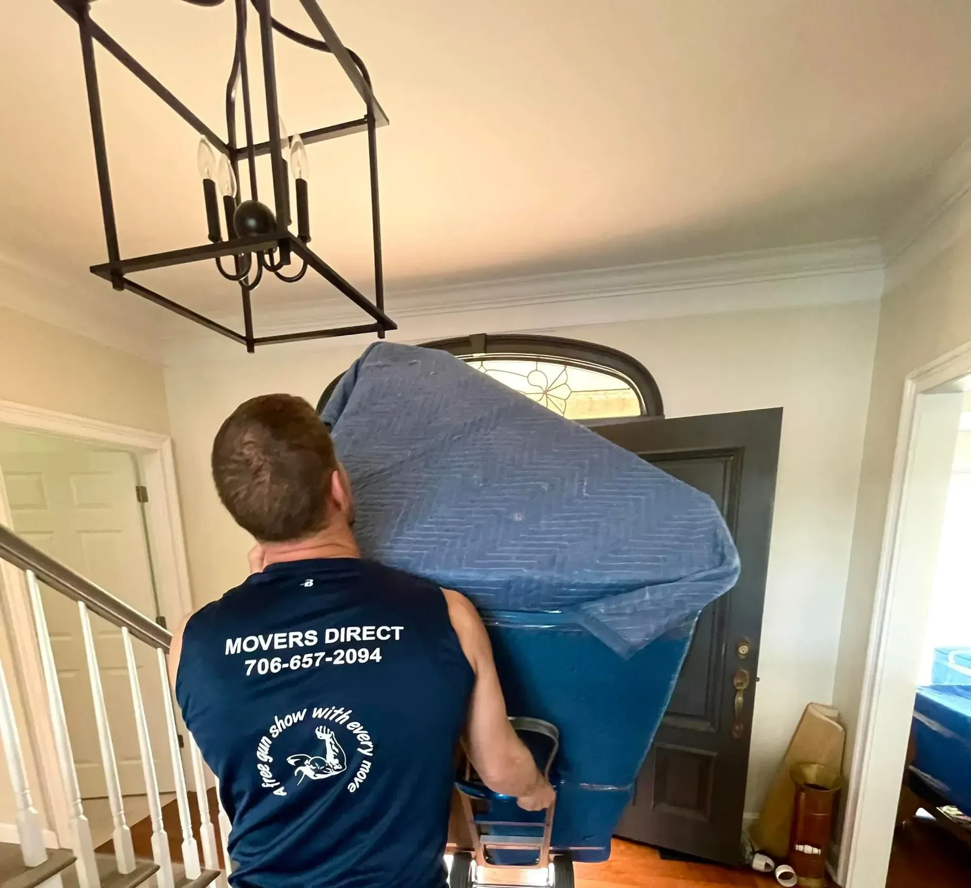 A person in a blue shirt from Movers Direct carries a large, blue, wrapped item through a doorway.