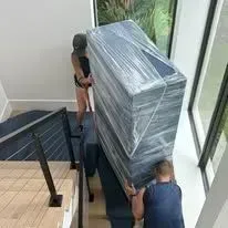 Two people carry a large, wrapped object up stairs. One person is at the top, one at the bottom.