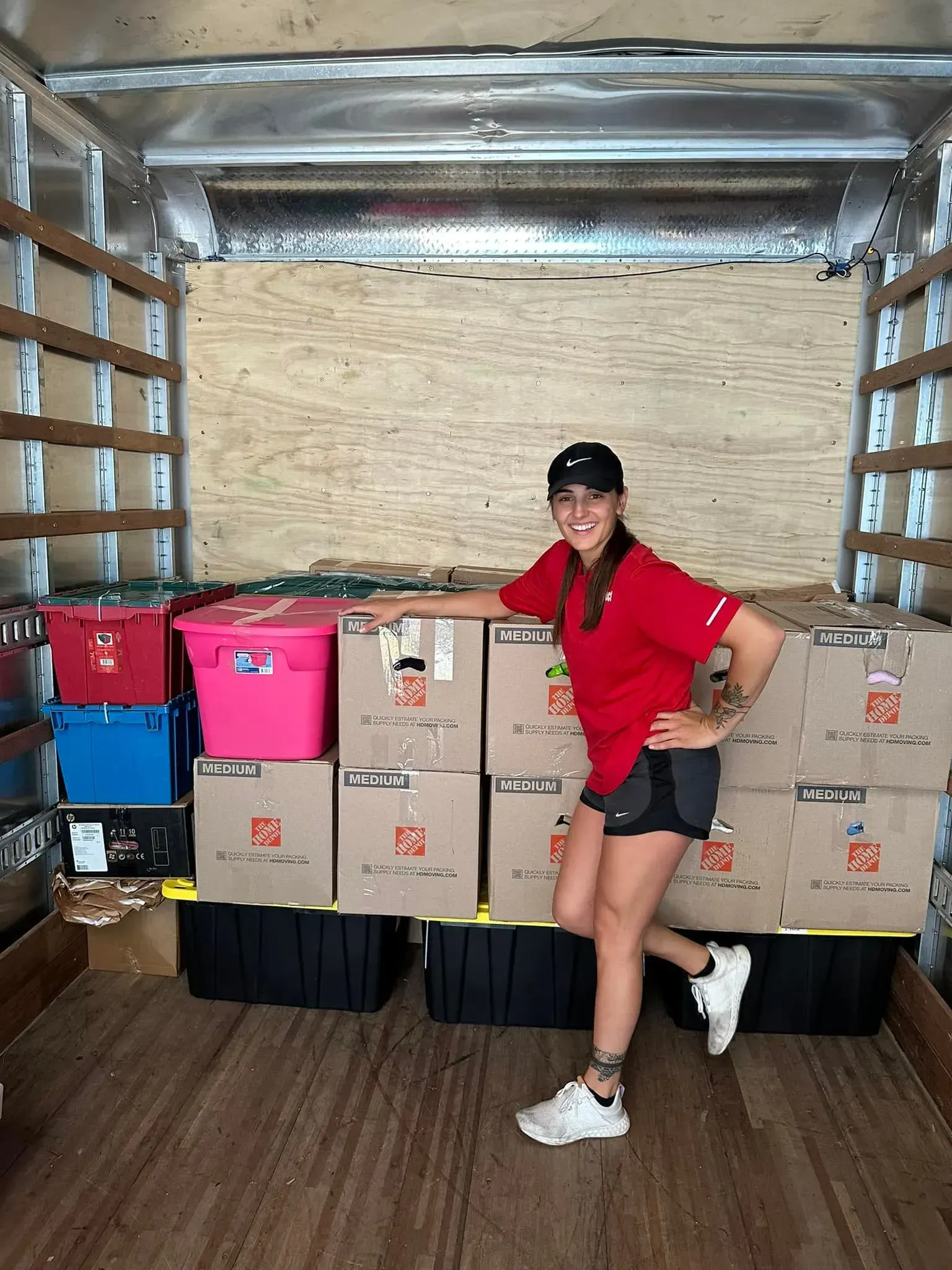 Woman in red shirt and shorts smiles in a moving truck, surrounded by packed boxes and bins.