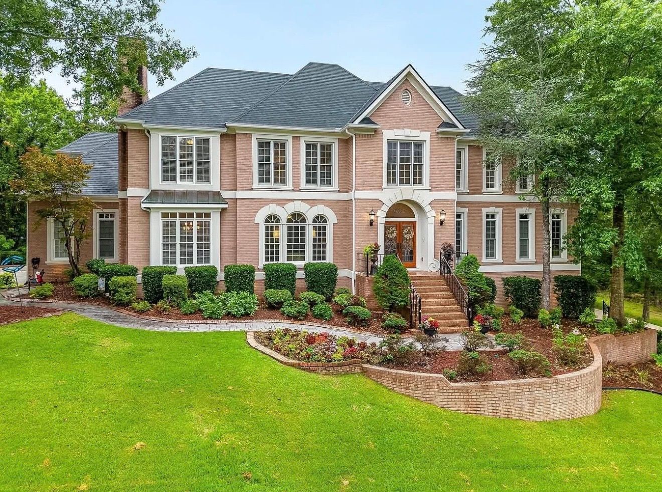 Two-story brick house with a dark roof and manicured lawn. Entrance features stairs and landscaping.