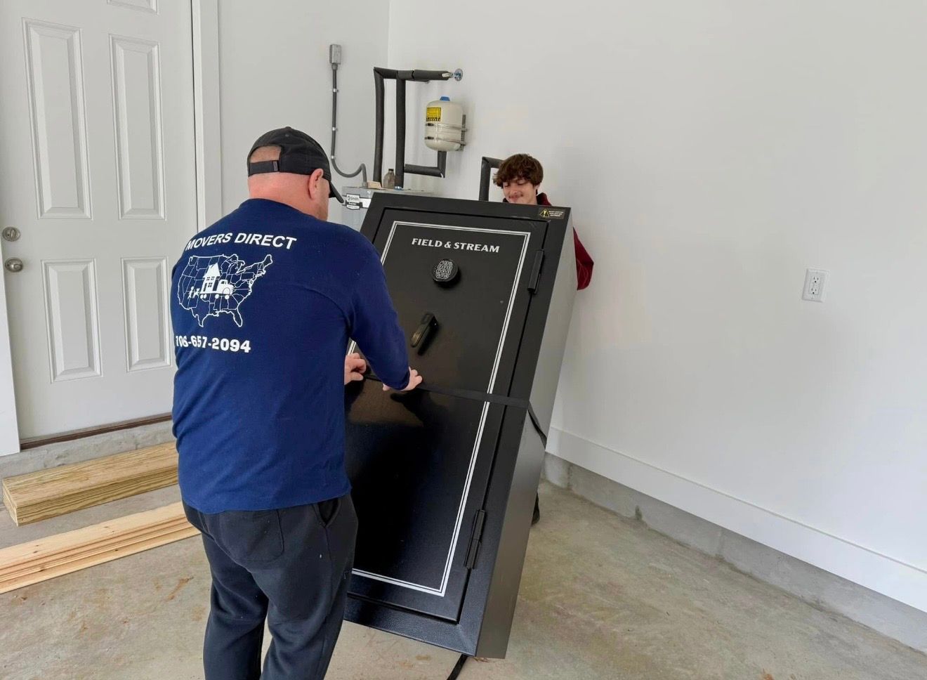 Two people lift a large, black safe in a garage. One man wears a blue shirt with a logo.