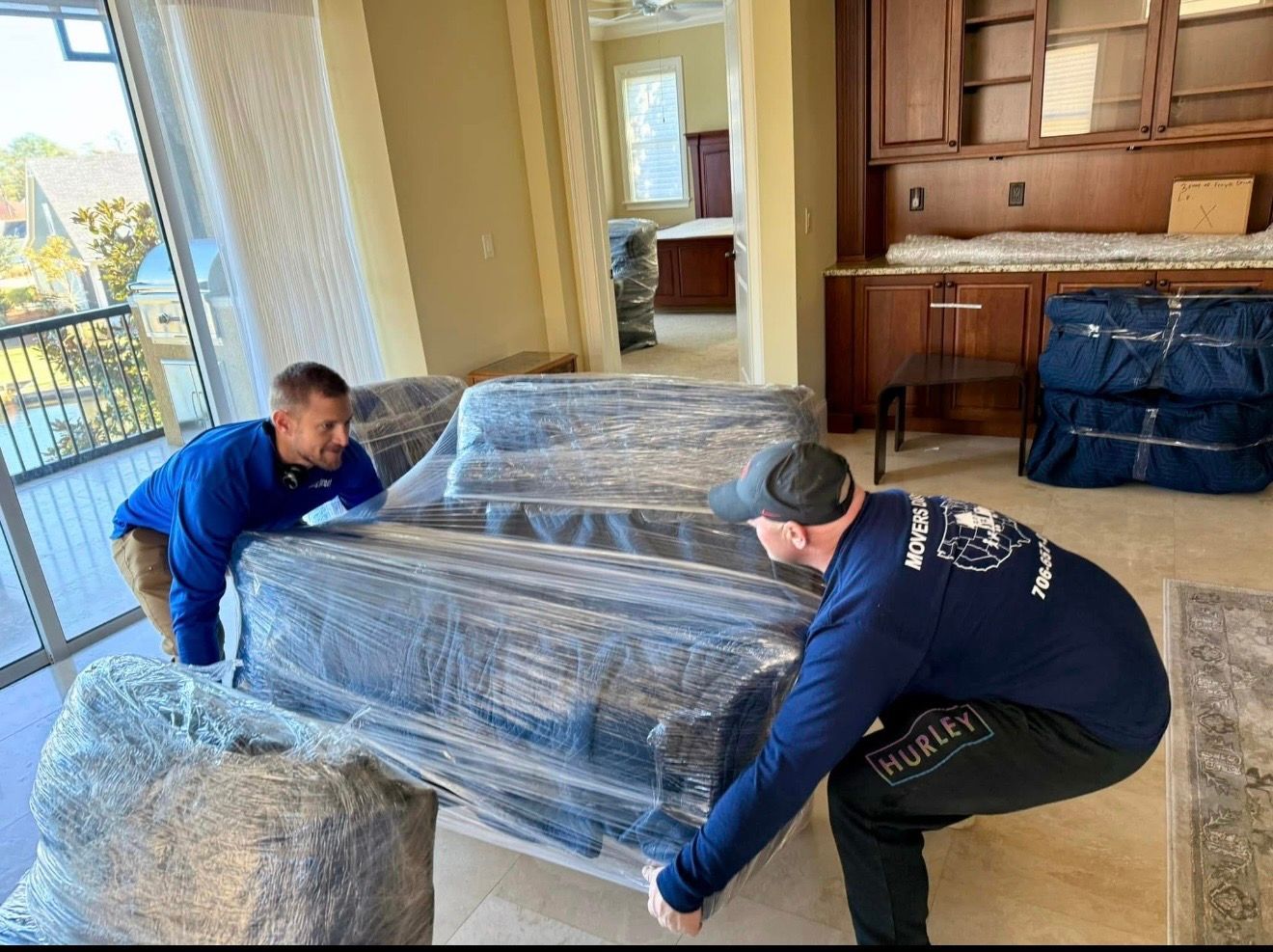 Two movers carrying a wrapped sofa in a home, near a balcony.