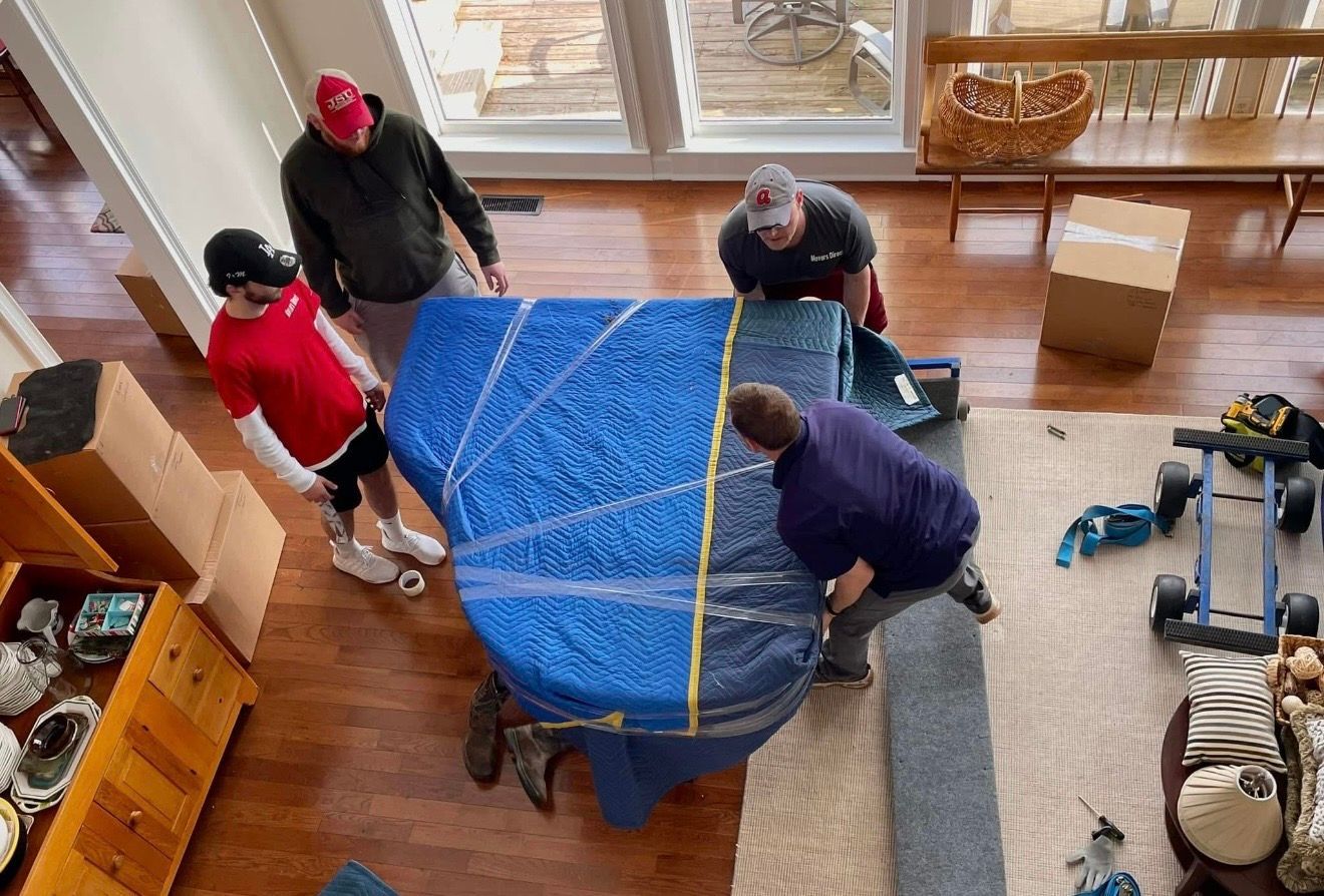 Movers in a living room, lifting a blue-wrapped piano. Other boxes and furniture are visible nearby.