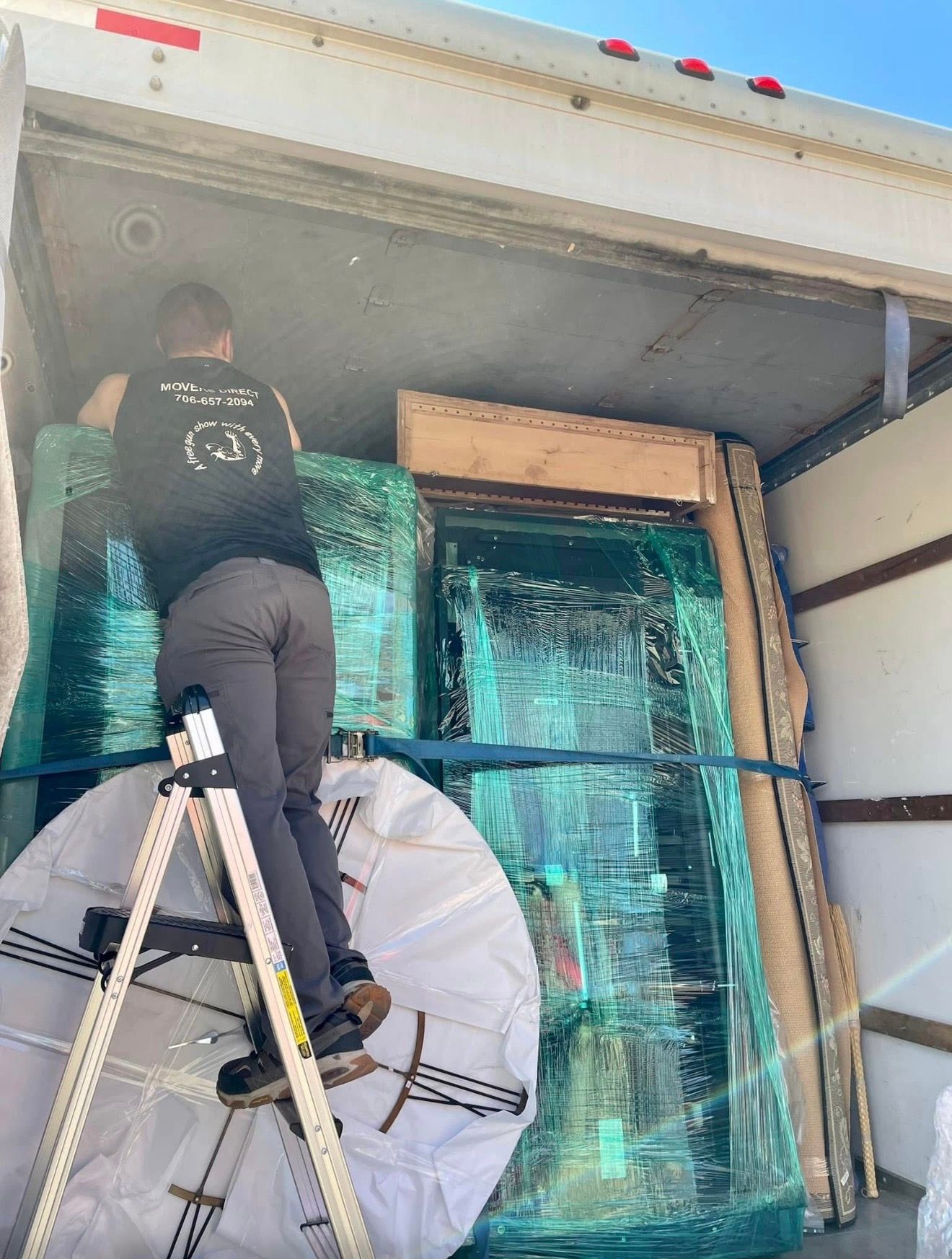 Person on a ladder loading large wrapped items, including a mirror and furniture, into a truck.