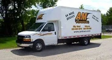 White AIM Towing box truck parked on a paved road next to grass and trees.