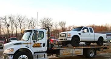 Tow truck hauling a white pickup truck. The tow truck is white with the AM logo on the door, outdoors.