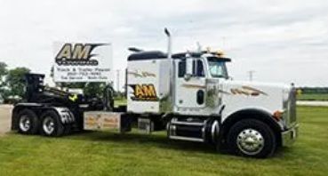 White semi-truck with AM Enterprises logo on a grassy field; truck and trailer with a flatbed.