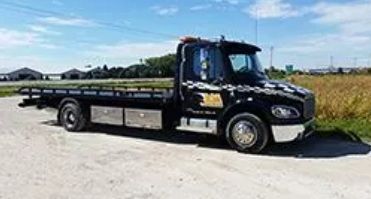 Black flatbed tow truck parked on a gravel road, sunny day.