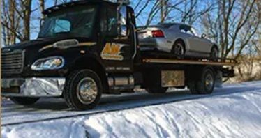 Black tow truck carrying a silver car on a snowy road.