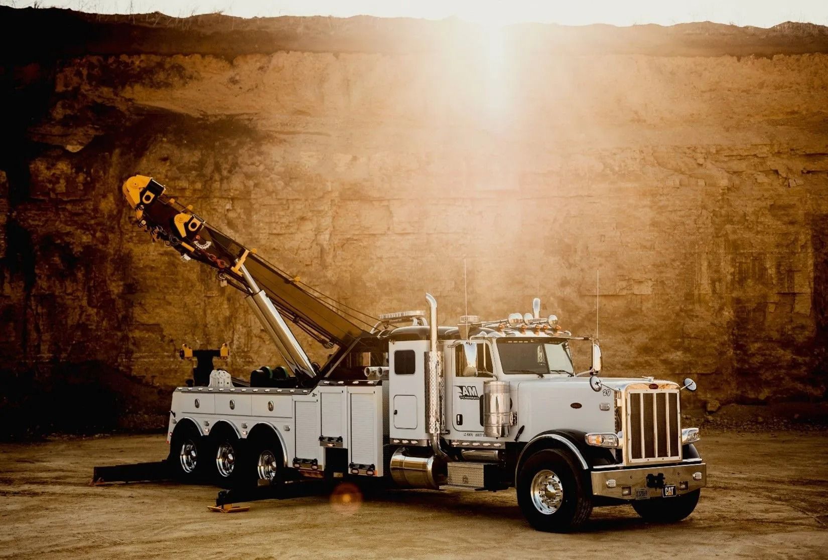 White tow truck with raised boom in front of a rock wall, with sunlight shining down.