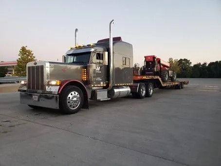 Chrome semi-truck hauling a tractor on a flatbed trailer, parked on asphalt in a parking lot.