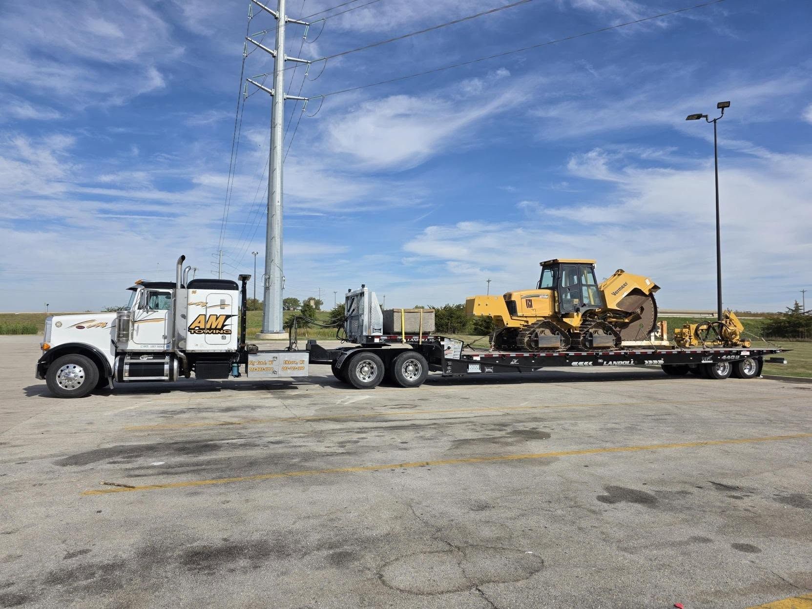 A white semi-truck hauls yellow construction equipment on a flatbed trailer in a parking lot.