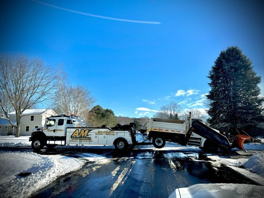 White truck towing a dump truck with snow plow on a snow-covered street under a blue sky.