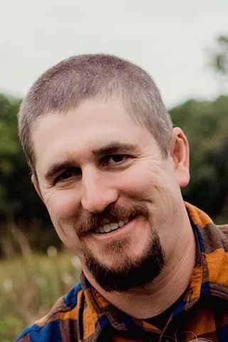 Man with short, graying hair and a goatee smiles outside, wearing a plaid shirt.