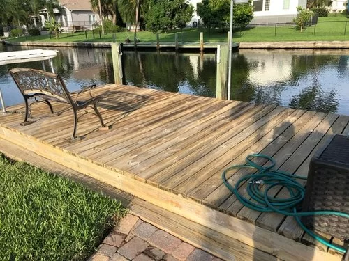 Wooden dock beside calm water with a chair, coiled hose, and grassy shoreline nearby.