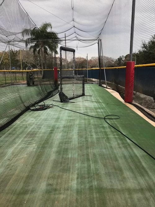 Outdoor cricket practice lane with green turf, netting, and red posts under a canopy.