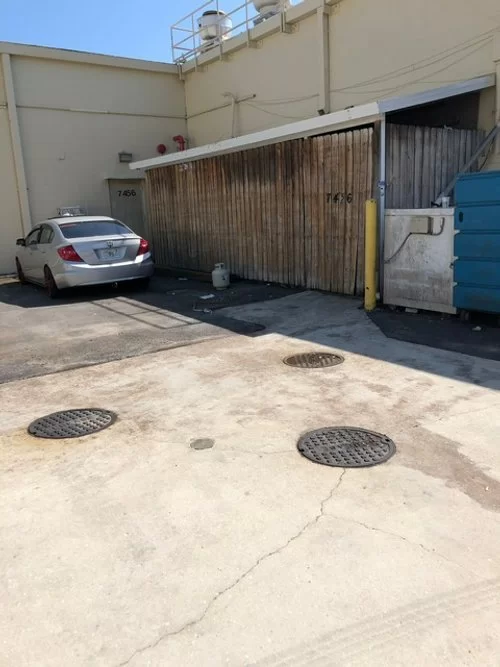 Industrial alley with parked silver car, corrugated shed, dumpsters, and concrete ground with manhole covers.