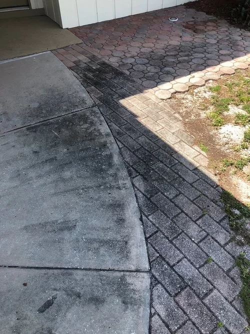 Sidewalk and driveway meeting at a rough, cracked brick path beside a house entrance.