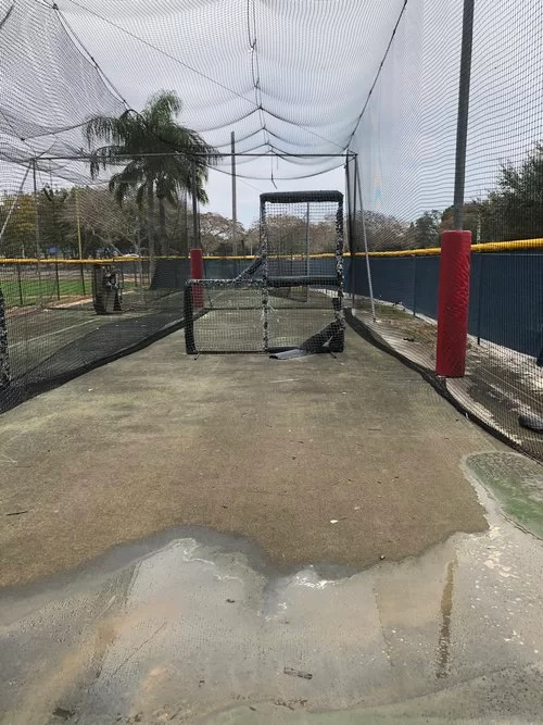 Outdoor batting cage with netting, puddled concrete, and a small practice gate in the center.