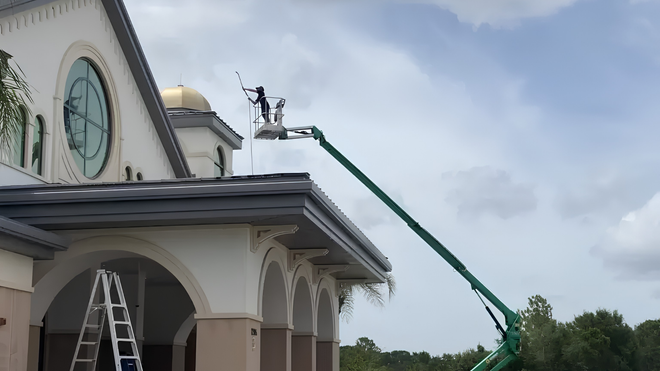 Workers in a green boom lift repair the roof of a building with arches and a clock face.