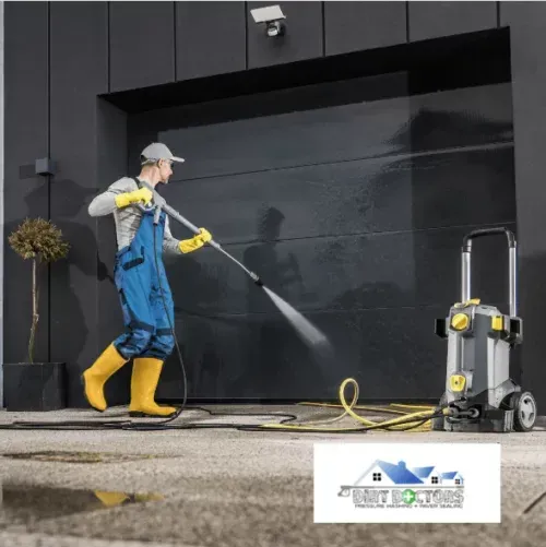 Worker in blue overalls pressure-washing a black wall beside a portable washer