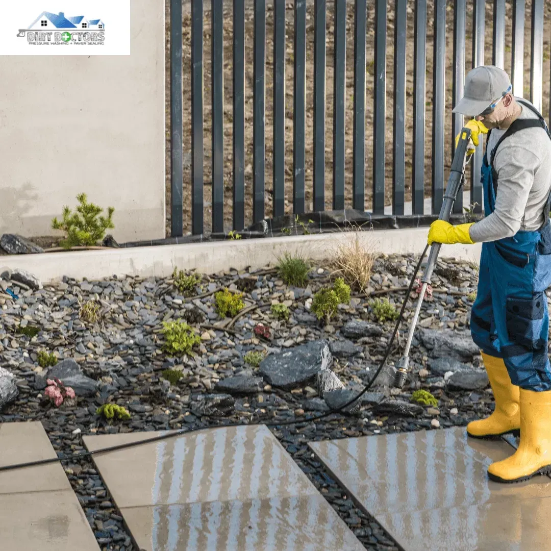 Worker in yellow boots pressure washing a sidewalk beside a fence and gravel bed.