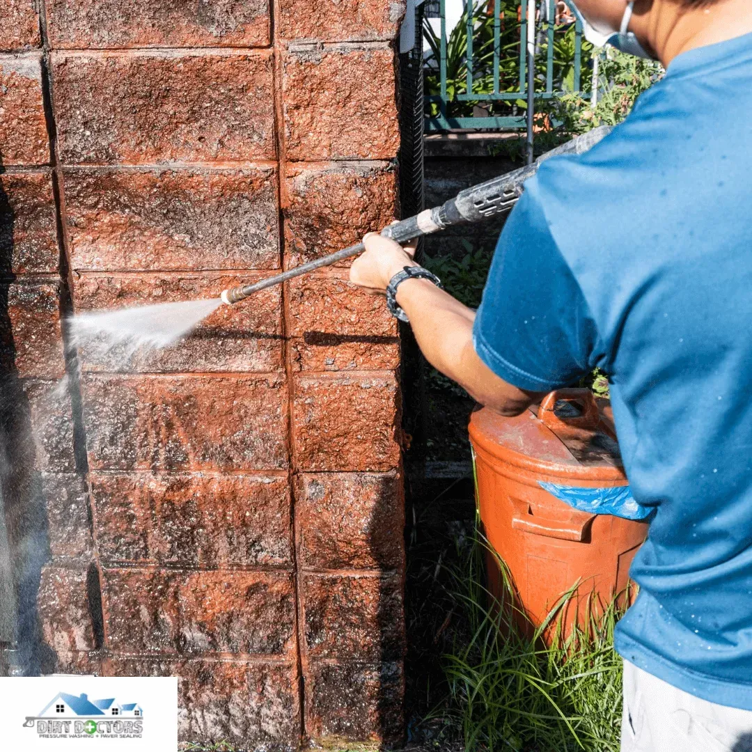 Person pressure-washing a red brick wall outdoors beside an orange tank