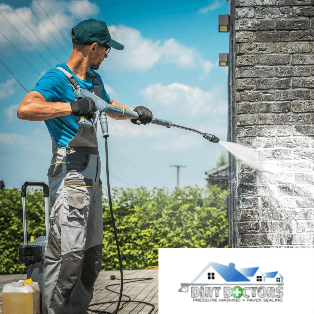 Worker pressure-washing a stone wall outdoors under a blue sky with a hedge in the background