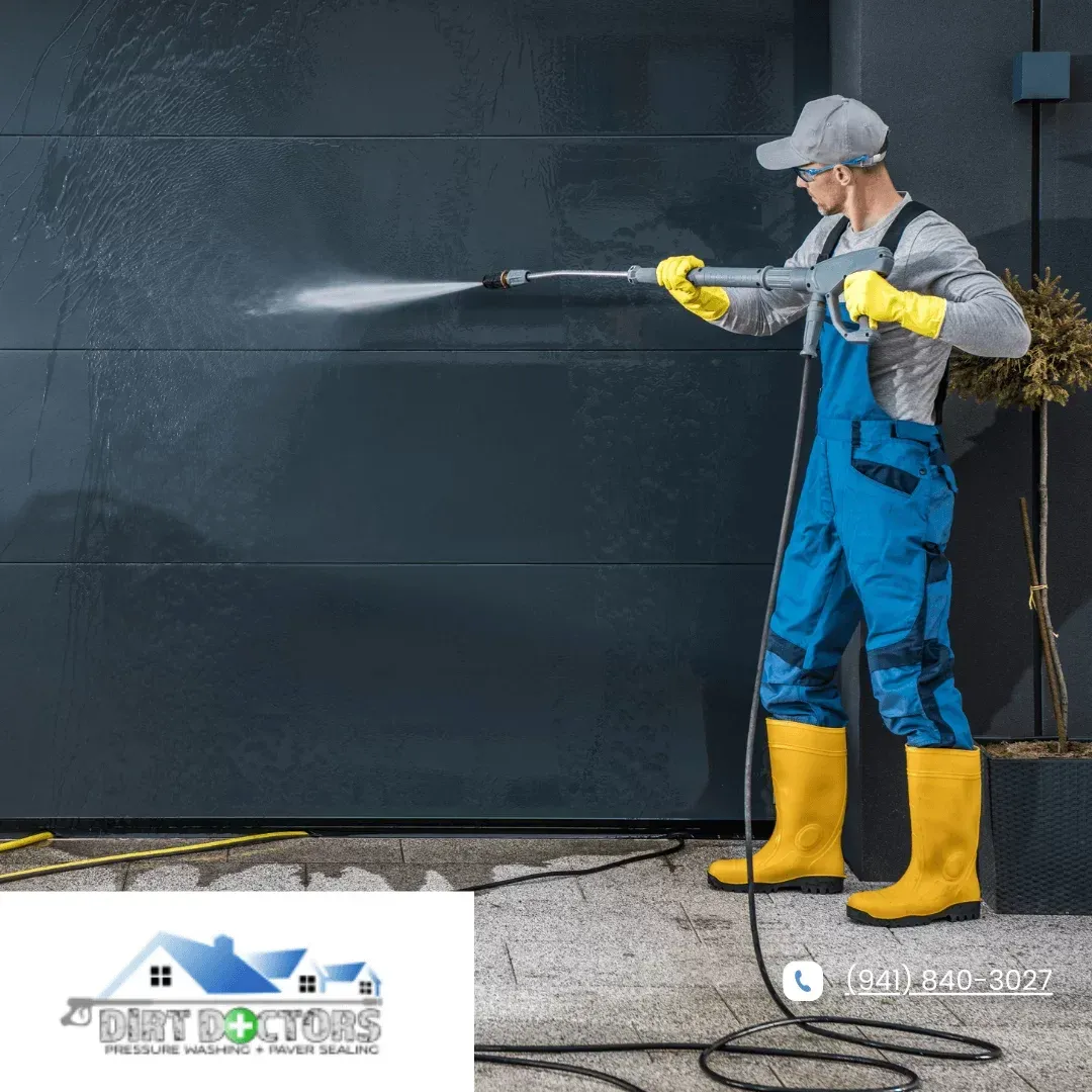 Worker in blue overalls pressure-washes a dark wall, wearing yellow gloves and boots.