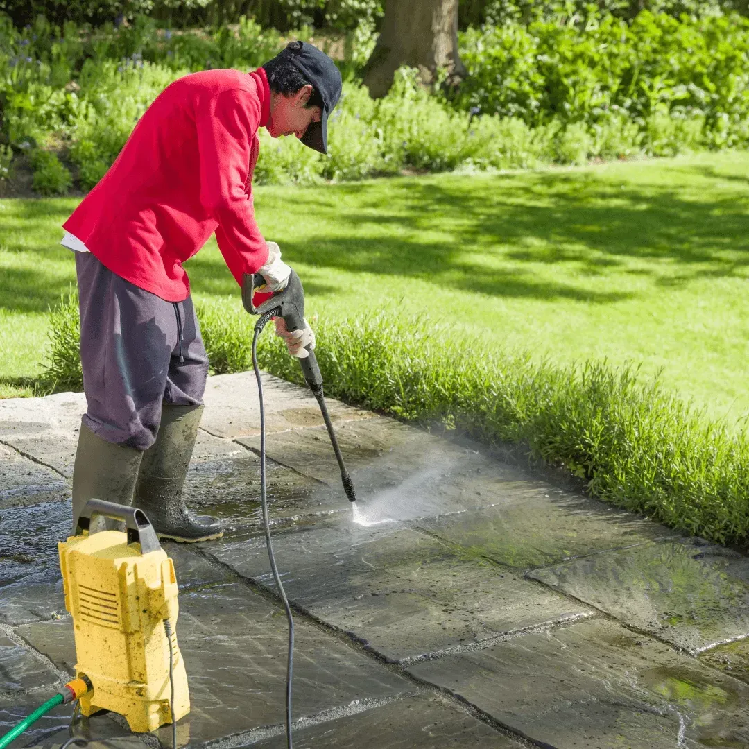 Person pressure-washing a stone patio beside a lawn, wearing a red shirt and boots.