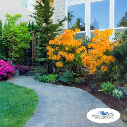 Curved garden path beside bright orange and pink flowering shrubs near a house with large windows