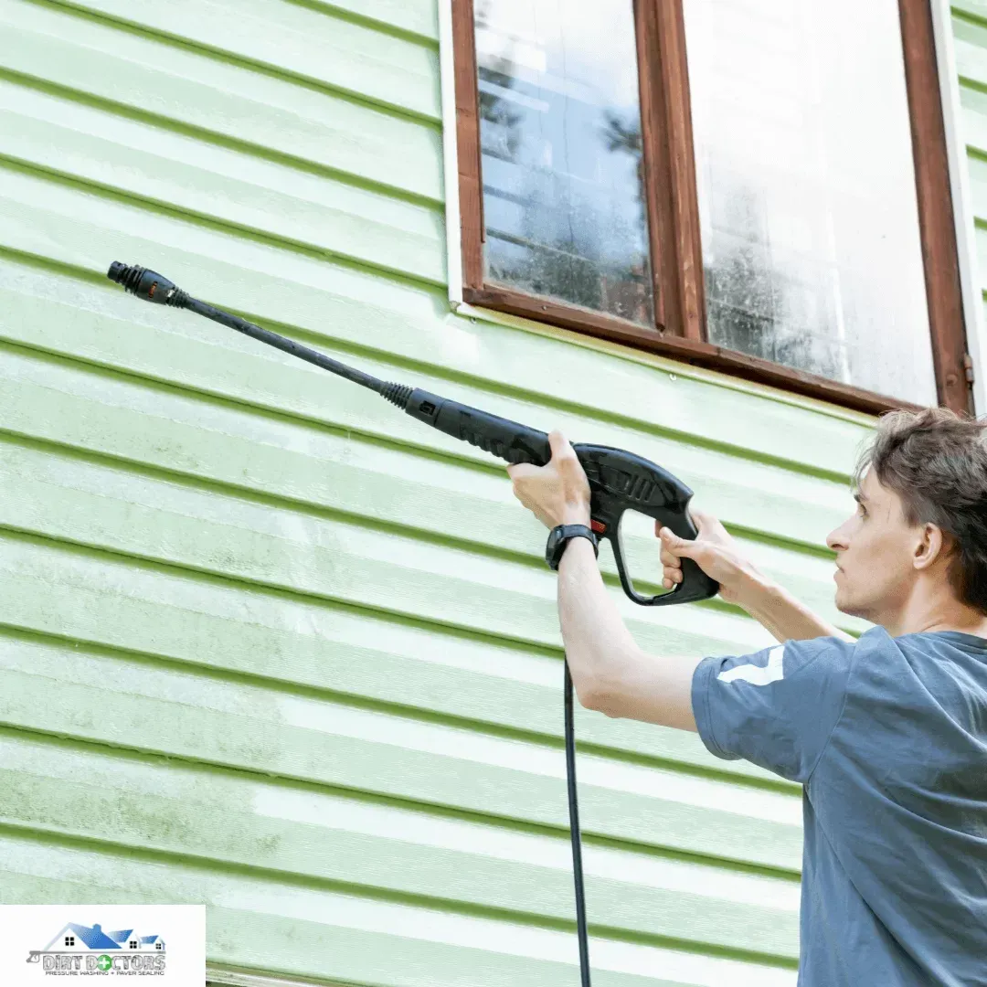 Person pressure-washing green house siding with a long black nozzle near a window