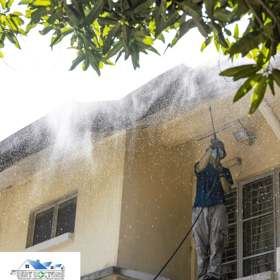 Person pressure-washing a beige building wall under tree branches, with water spraying from the roofline.