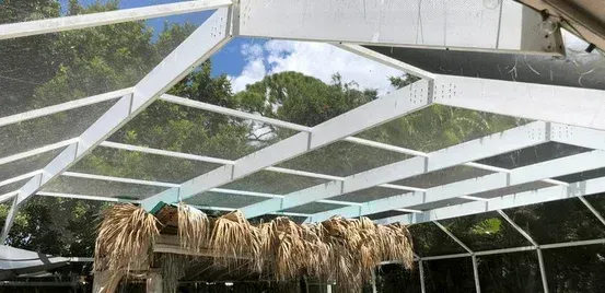 White greenhouse roof frame with hanging dried plants beneath, against a backdrop of trees and sky