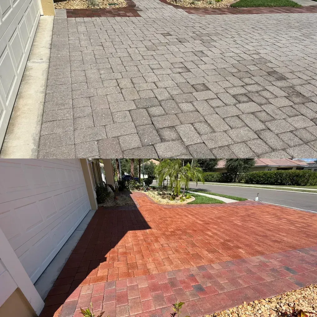Paved driveway and red brick patio beside a white house, with a road and greenery beyond.