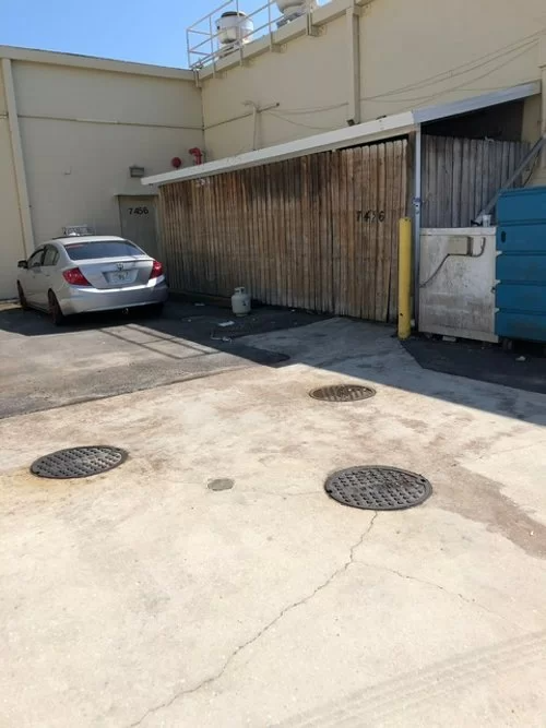 Parking lot with a silver car beside a wooden shed and blue dumpster against a beige wall.