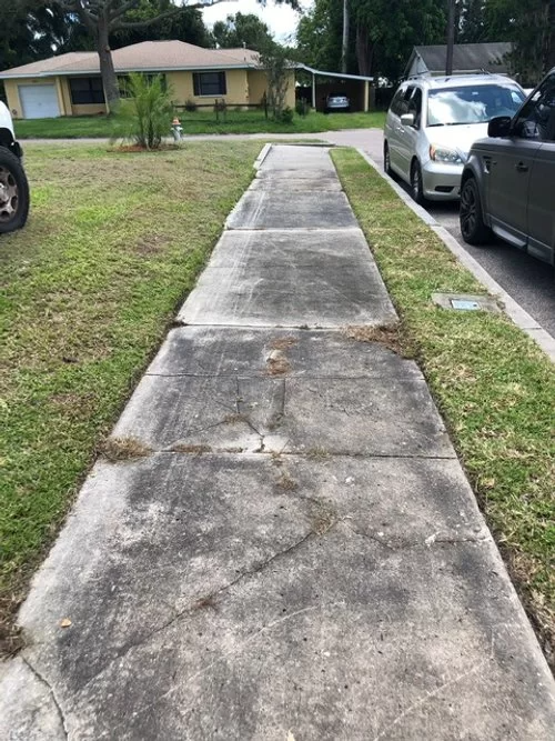 Cracked concrete sidewalk leading to a house, with parked cars along a grassy driveway.