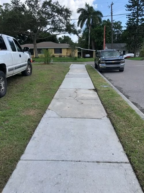 Cracked sidewalk beside a residential street with parked vehicles and houses in the background