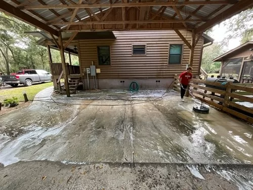 Covered concrete patio being power washed by a person in a red shirt and white hat