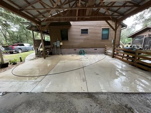 Covered concrete driveway beside a wooden house, wet from rain, with a hose and parked cars nearby