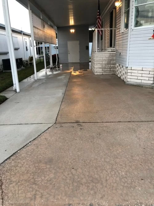 Covered concrete walkway beside houses, with white siding, a porch, and an American flag.