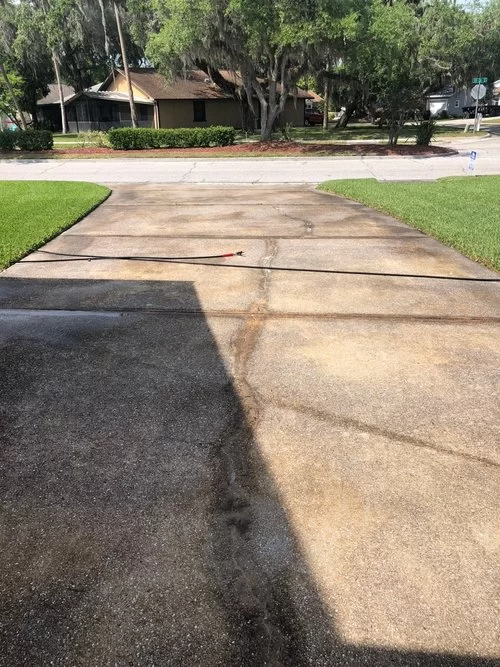 Driveway with dark and light pavement patches, leading to a street and trees in a suburban neighborhood.