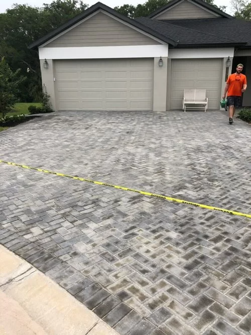 Driveway with gray pavers leading to a garage; a person in an orange shirt stands at the right edge.